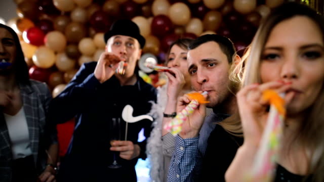 Group of friends at a private party. They are holding drinks, cocktails and wine, and blowing into party noise makers.