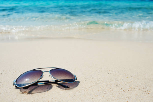istock Sunglasses on white sand on blue wave background. Vacation by the warm sea 1200987480