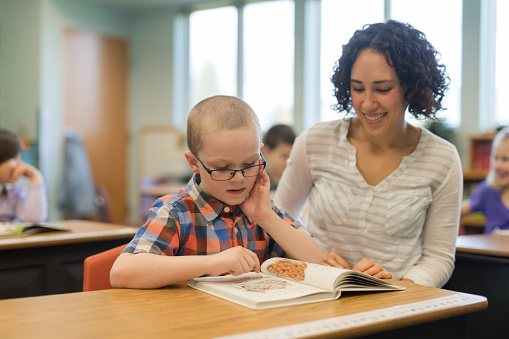 An ethnic female teacher helps a young elementary boy work on a spelling assignment at his desk. He is turning through the pages and sounding out words.