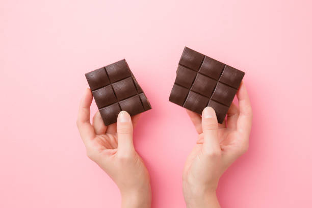 istock Young woman hands holding broken chocolate bar on light pink table background. Pastel color. Sweet snack. Closeup. Point of view shot. Top down view. Two sides. 1201003853