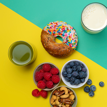 This is an overhead colorful photo of a split diagonal image showing half of a donut and half of a bagel seamlessly joined together. One half of the side is showing healthy food and the other half of the side is showing junk food