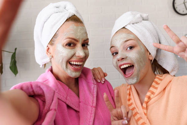 madre y su hija tomando selfie en el teléfono inteligente con máscara facial, albornoz y toalla en la cabeza - facial mask spa treatment cucumber human face fotografías e imágenes de stock