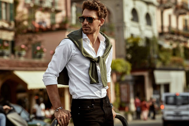 Stylish man wearing sunglasses and white shirt. City life Stylish man wearing sunglasses and white shirt with tied sweater on shoulders cool attitude fashion stock pictures, royalty-free photos & images