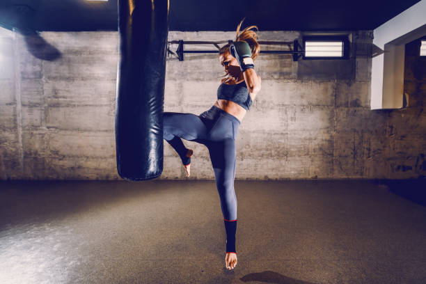 joven ajuste atractivo musculoso mujer kick boxer en ropa deportiva con guantes de boxeo kicking bolsa de boxeo con rodilla. - boxing womens fotografías e imágenes de stock