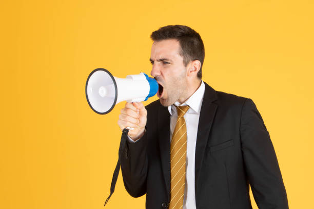 istock Portrait of a handsome businessman wearing white suit and tie while using megaphone, screaming with yellow background. 1201083053