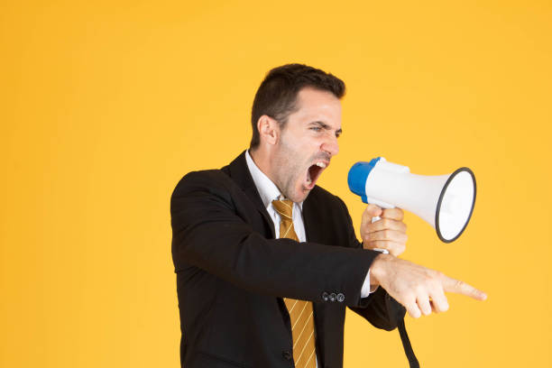 istock Portrait of a handsome businessman wearing white suit and tie while using megaphone, screaming with yellow background. 1201083067
