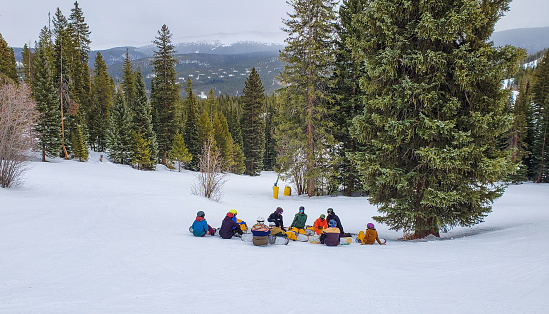 Group of snowboarders sitting on snow in Colorado ski resort in winter; pines and spruce trees and mountain range in background