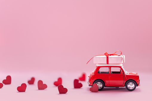 Red toy car delivering carrying on roof gift box with small red hearts on pink background. Valentine's day concept. Banner