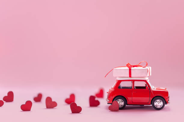 Red toy car delivering carrying on roof gift box with small red hearts on pink background. Valentine's day concept. stock photo