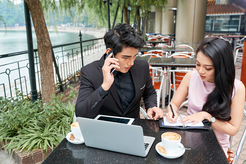 Horizontal shot of young Asian man and woman sitting together in modern cafe working together on project