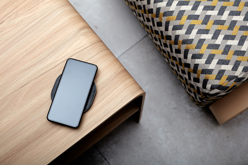 A mobile phone charging wirelessly is on a wooden table，Vertical shooting of mobile phone wireless charging scene, background with wood desktop and cloth sofa,