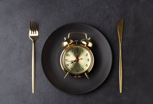 Golden alarm clock on a black plate with a golden knife and fork Golden alarm clock on a black plate with a golden knife and fork. Intermittent fasting concept. Horizontal orientation, top view. fasting stock pictures, royalty-free photos & images