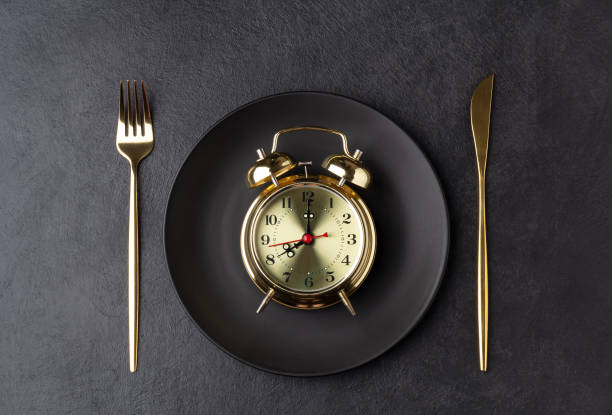 Golden alarm clock on a black plate with a golden knife and fork Golden alarm clock on a black plate with a golden knife and fork. Intermittent fasting concept. Horizontal orientation, top view. Regimen stock pictures, royalty-free photos & images