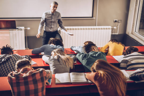 Large group of exhausted students sleeping at lecture hall. Large group of tired college students fell asleep on a class at lecture hall while their teacher is in disbelief. male college student sleeping through lecture stock pictures, royalty-free photos & images