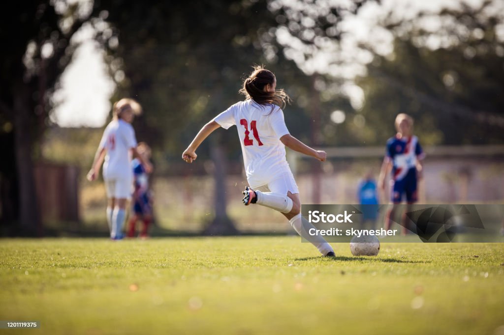 Rear view of determined female soccer player kicking the ball on a match. Back view of female soccer player kicking the ball during a match on a stadium. Soccer Stock Photo