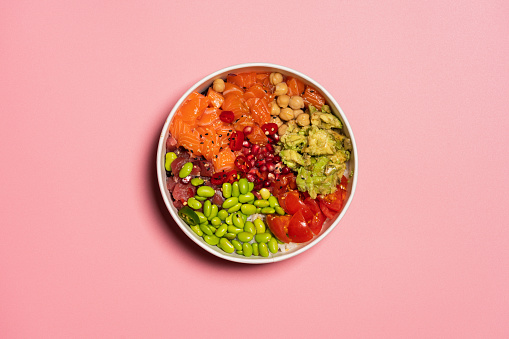 Hawaiian poke bowl with rice, salmon, avocado, tomatoes, tuna, chickpeas, pomegranate and edamame. Top view from above. Pink background for copy space.