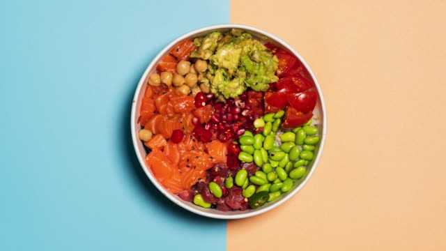Stop motion video of an hawaiian poke bowl with rice, salmon, avocado, tomatoes, tuna, chickpeas, pomegranate and edamame. Top view from above. Backgrounds of multiple colors changing for copy space.