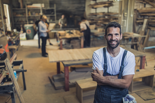 Young happy carpenter standing in a workshop with his arms crossed and looking at camera. Thee are people in the background.