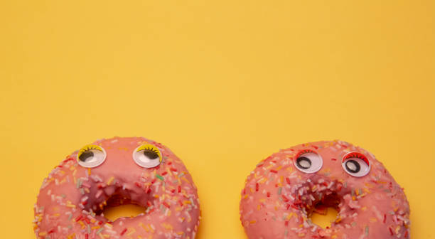 istock Two glazed pink donuts isolated with funny faces on yellow background. Conceptual image 1201162080