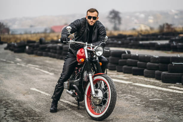 istock Bearded man in sunglasses and leather jacket looking at the camera while sitting on a motorcycle on the road. Behind him is a row of tires 1201162540