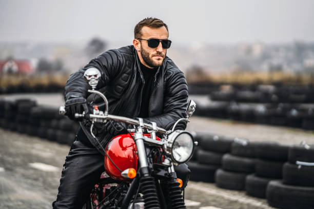 istock Bearded man in sunglasses and leather jacket looking at the camera while sitting on a motorcycle on the road. Behind him is a row of tires 1201162565