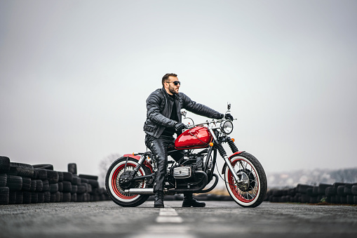 Red motorbike with rider. A man in a black leather jacket and pants stands sideways in the middle of the road. Tires are laid on the background.