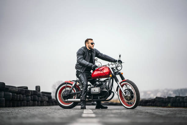 Red motorbike with rider. A man in a black leather jacket and pants stands sideways in the middle of the road. Tires are laid on the background stock photo