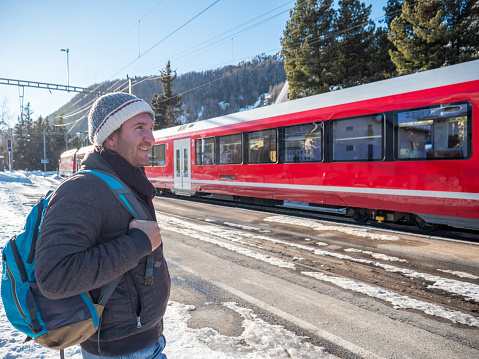 Tourist man waiting for his train on railroad platform in winter. Red train in Switzerland, Graubunden Canton. Eco tourism travel vacations concept