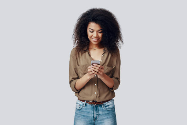 istock Beautiful young African woman using smart phone and smiling while standing against grey background 1201179304