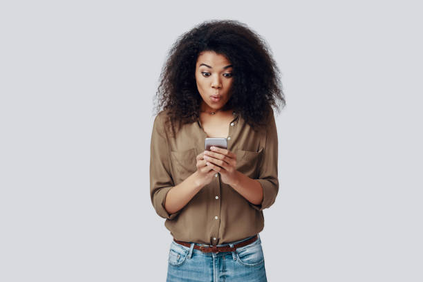 istock Surprised young African woman using smart phone and making a face while standing against grey background 1201179405