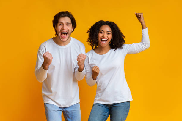 Overjoyed multiracial couple celebrating success with raised fists, exclaiming with excitement We Did It. Overjoyed multiracial couple celebrating success with raised fists, emotionally reacting to victory, posing together over yellow background clingy girlfriend stock pictures, royalty-free photos & images