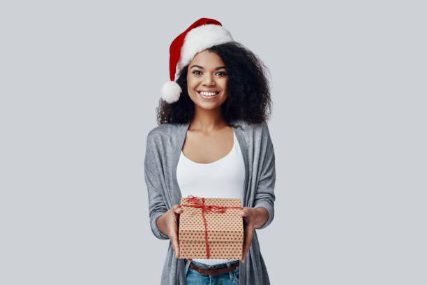 belle jeune femme africaine dans le chapeau de santa te donnant le cadeau et le sourire de noel tout en restant contre le fond gris - beautiful women gift christmas photos et images de collection