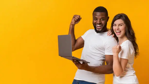 Photo of Joyful interracial couple holding laptop, celebrating success and exclaiming in excitement