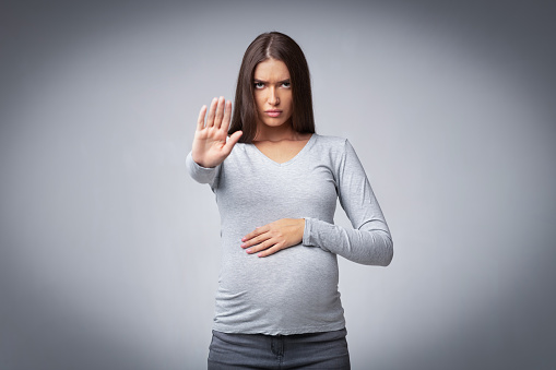 Pregnant woman gesturing stop looking severely at camera forbidding or warning posing in studio over gray background. Selective focus