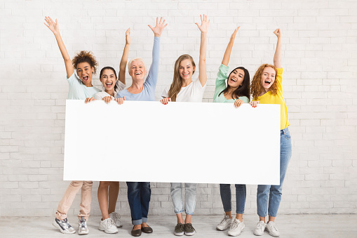 Group Of Cheerful Women Holding Blank White Poster Waving Hello Posing Near Brick Wall Indoor. Mockup