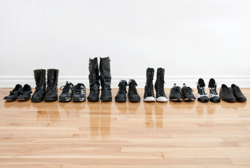 Row of black shoes and boots on a wooden floor, in front of a white wall.