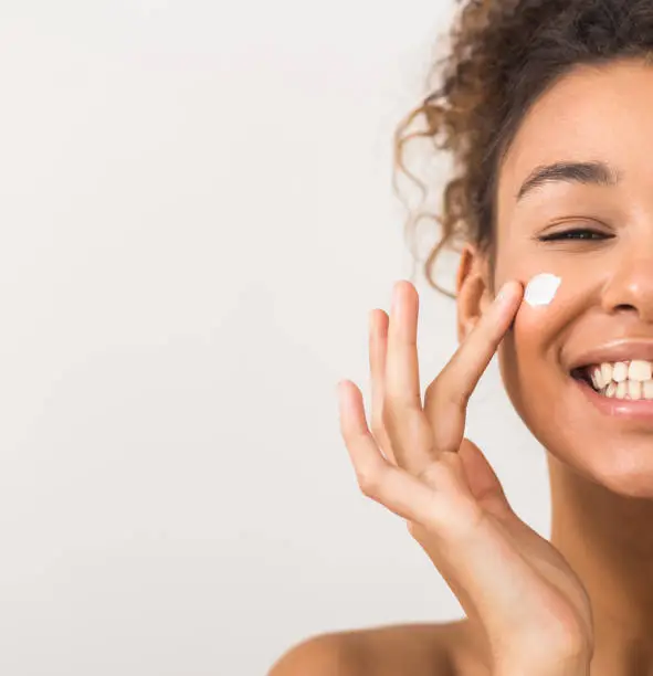 Photo of Face care. Happy black woman applying moisturizer cream on cheek