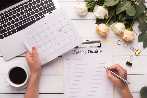 Bride Planning wedding writing checklist and choosing date in calendar, wooden background with cream colored roses and laptop