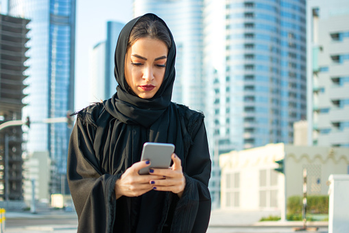 Portrait of beautiful Arab businesswoman wearing hijab using cell phone on the street with the skyscrapers in the background