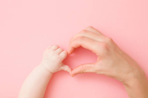 istock Heart shape created from infant and young mother hands on light pink table background. Pastel color. Lovely emotional, sentimental moment. Love, happiness and safety concept. Closeup. Top down view. 1201208384