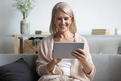 Smiling mature woman sit relax on sofa in living room enjoy watching movie on tablet at home, happy mature old female retiree use electronic gadget browse internet, read news, easy technology concept