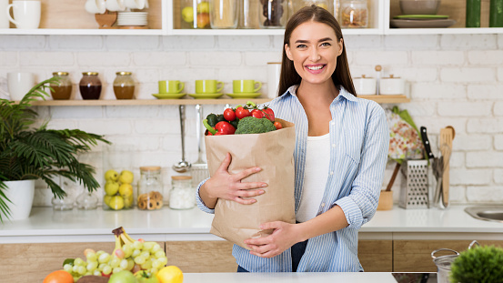 Housewife holding bag with vegetables after shopping in grocery. Woman standing in kitchen and smiling, panorama