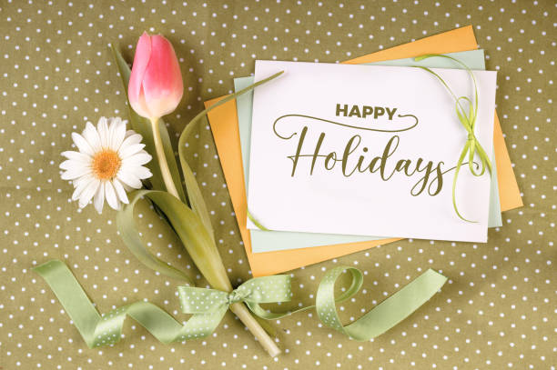 istock Greeting card with caption Happy Holidays on stack of paper cards decorated with Springtime flowers, daisy and tulip, and ribbons with bow on olive green table cloth with polka dots 1201218554