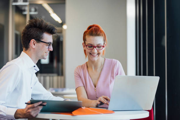 University Student on Work Placement Young man and woman sitting at a table working together with a laptop open in front of them. They are smiling two heads are better than one stock pictures, royalty-free photos & images