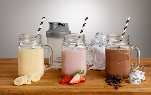 Banana, Strawberry and Chocolate milkshakes in Mason Jar glasses, with paper straws, ingredients and shaker on a wooden work top