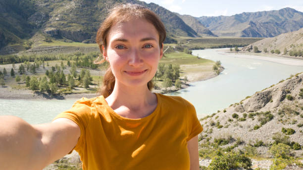 Brunette girl with a ponytail. Takes a selfie on the background of the river. Summer trip in nature. A trip to the Altai stock photo
