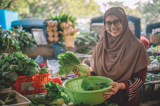 an asian malay female with eyeglasses and hijab looking at camera with her vegetable and basket at the vegetable stall smiling