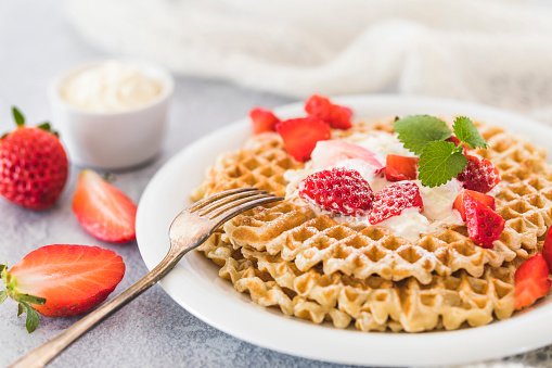 Swedish waffles with strawberries and whipped cream in a small bowl in the background. Next to the plate is a fork and whole and halved strawberries.