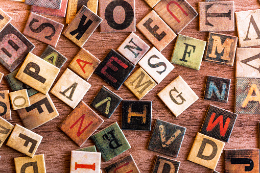 Color image depicting an overhead view of a large selection of colored alphabet letter tiles scattered, apparently haphazardly, across a wooden surface.