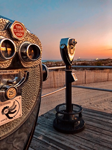 Coin operated binoculars on the boardwalk facing the beach and the pier at sunset with a glowing, warm peach sky